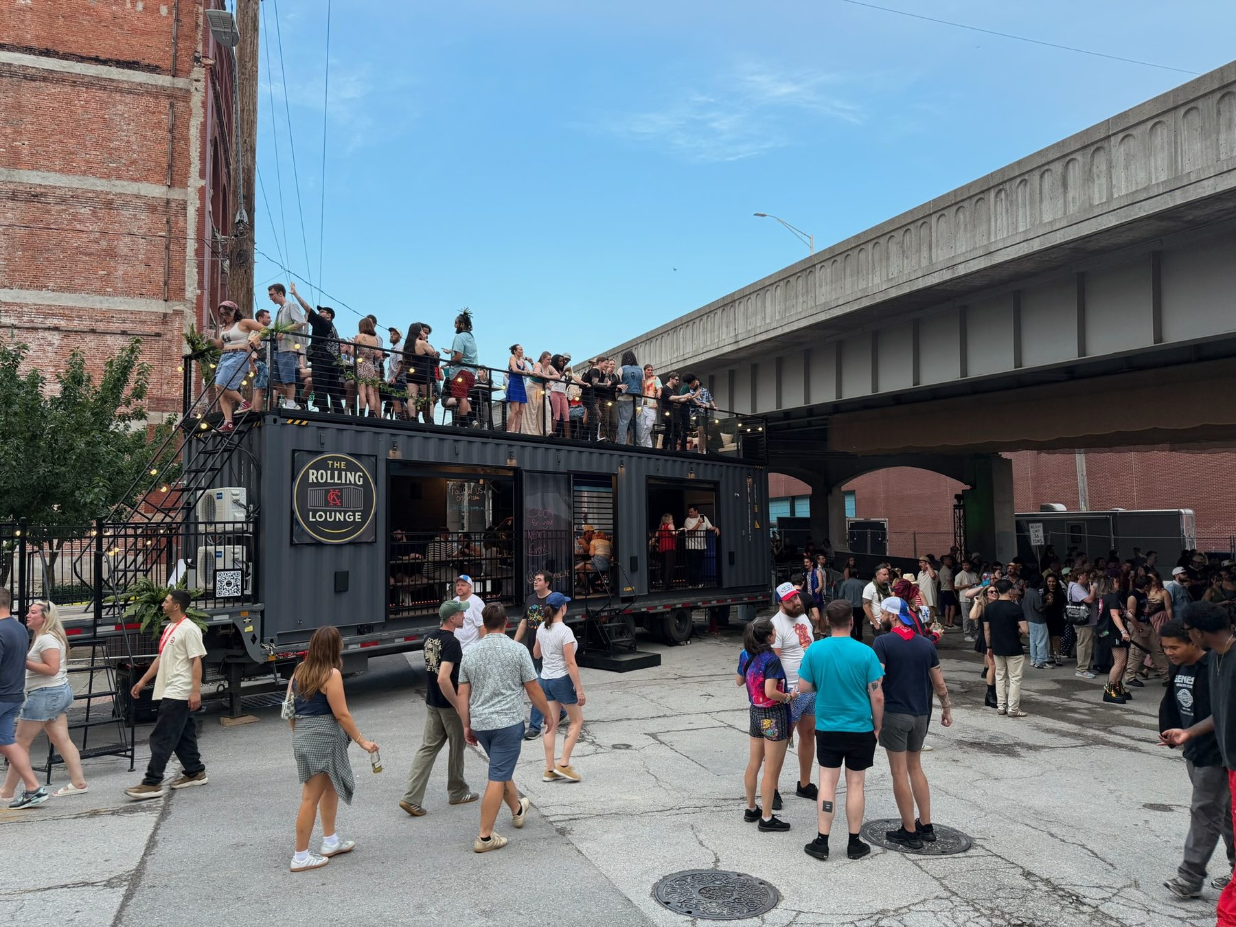 The Rolling Lounge parked under an overpass with a crowd on the rooftop and street