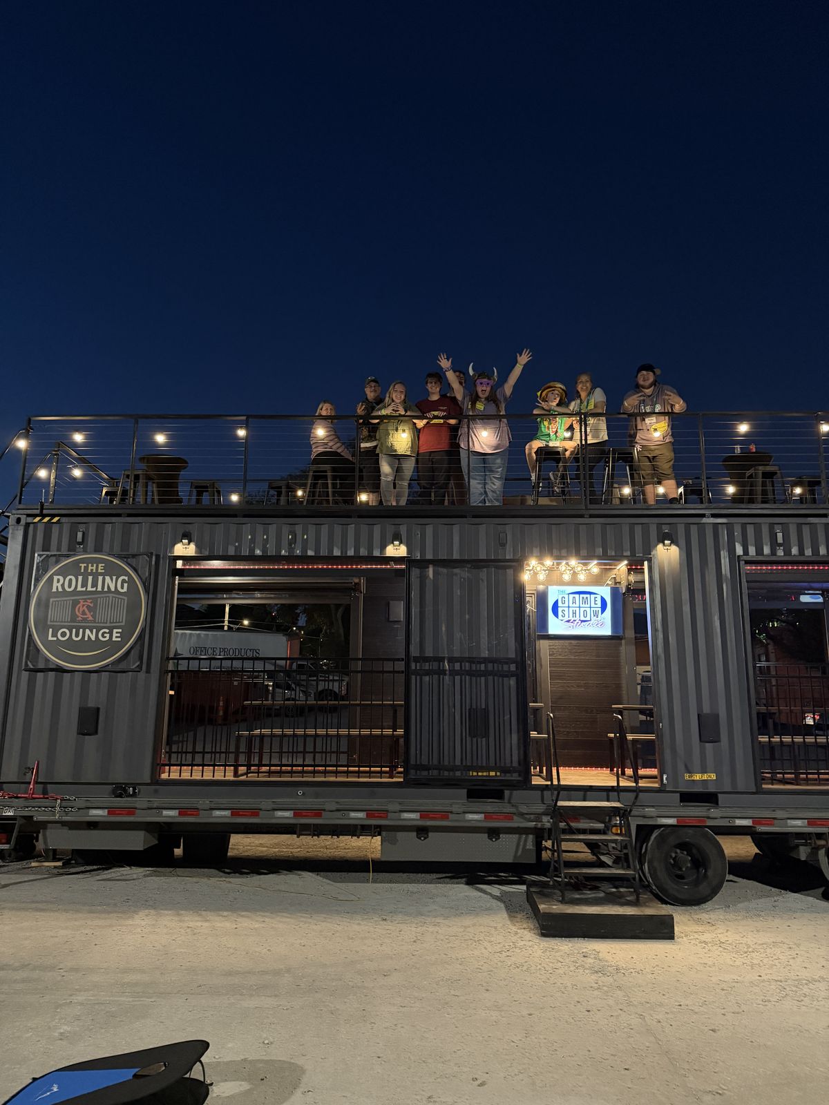 Guests on the Rolling Lounge rooftop at night during Game Show Studio, with a lit stage behind the deck